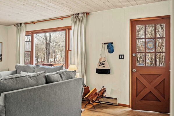 Una sala de estar acogedora con un sofá gris, una puerta de madera con paneles de vidrio y vistas a los árboles a través del ventanal.