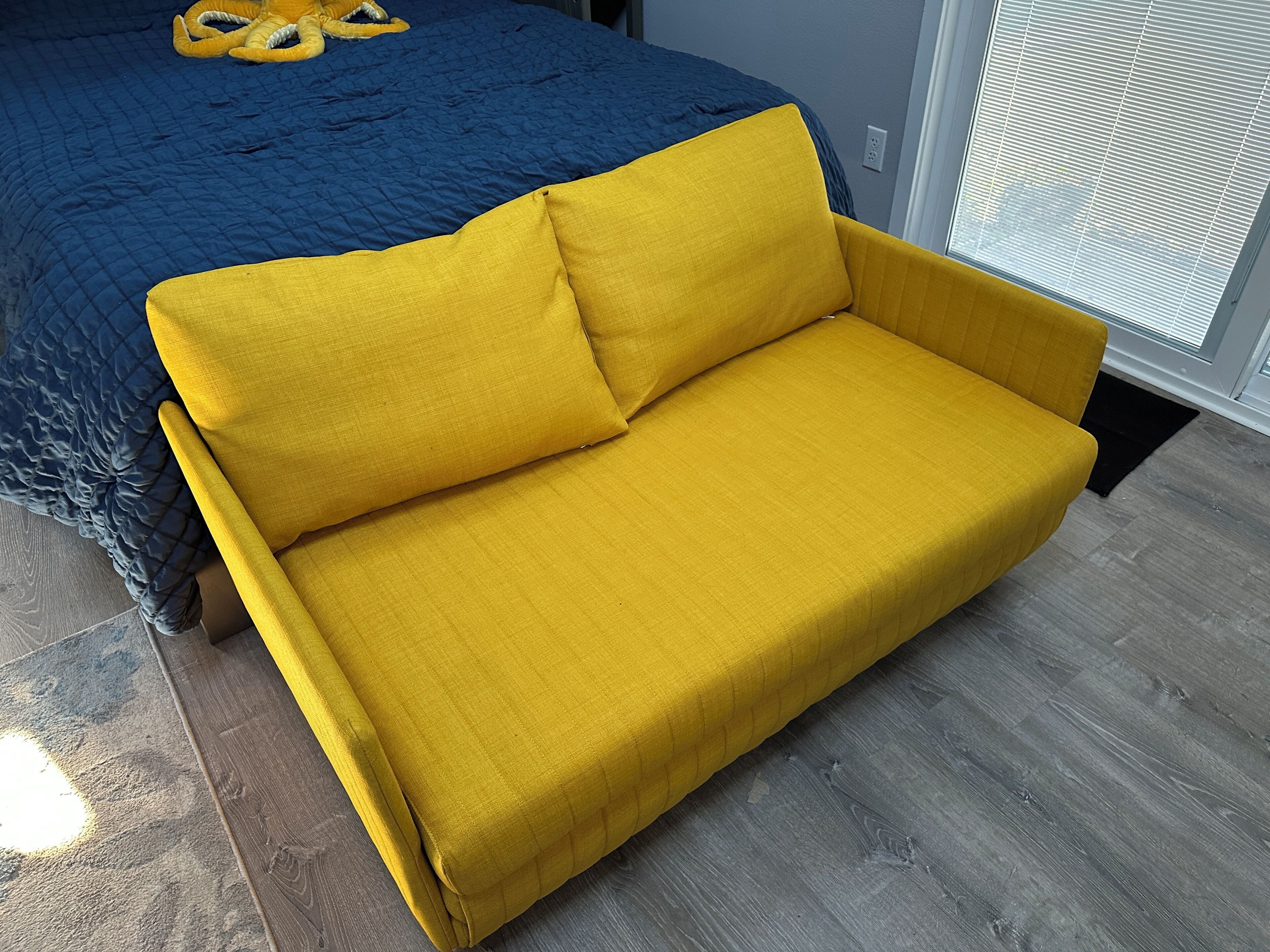 A yellow sofa with a blue quilted bedspread in the background.