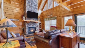 A log cabin interior with a stone fireplace, leather furniture, and a television mounted above it. - Anne's Place by Avantstay Cozy Cabin With Hot Tub, Games, Near Dollywood Attractions (Sevierville)