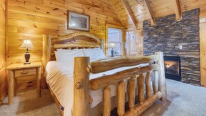 A log cabin bedroom with a stone fireplace, wooden bed, and a ceiling fan. - Anne's Place by Avantstay Cozy Cabin With Hot Tub, Games, Near Dollywood Attractions (Sevierville)