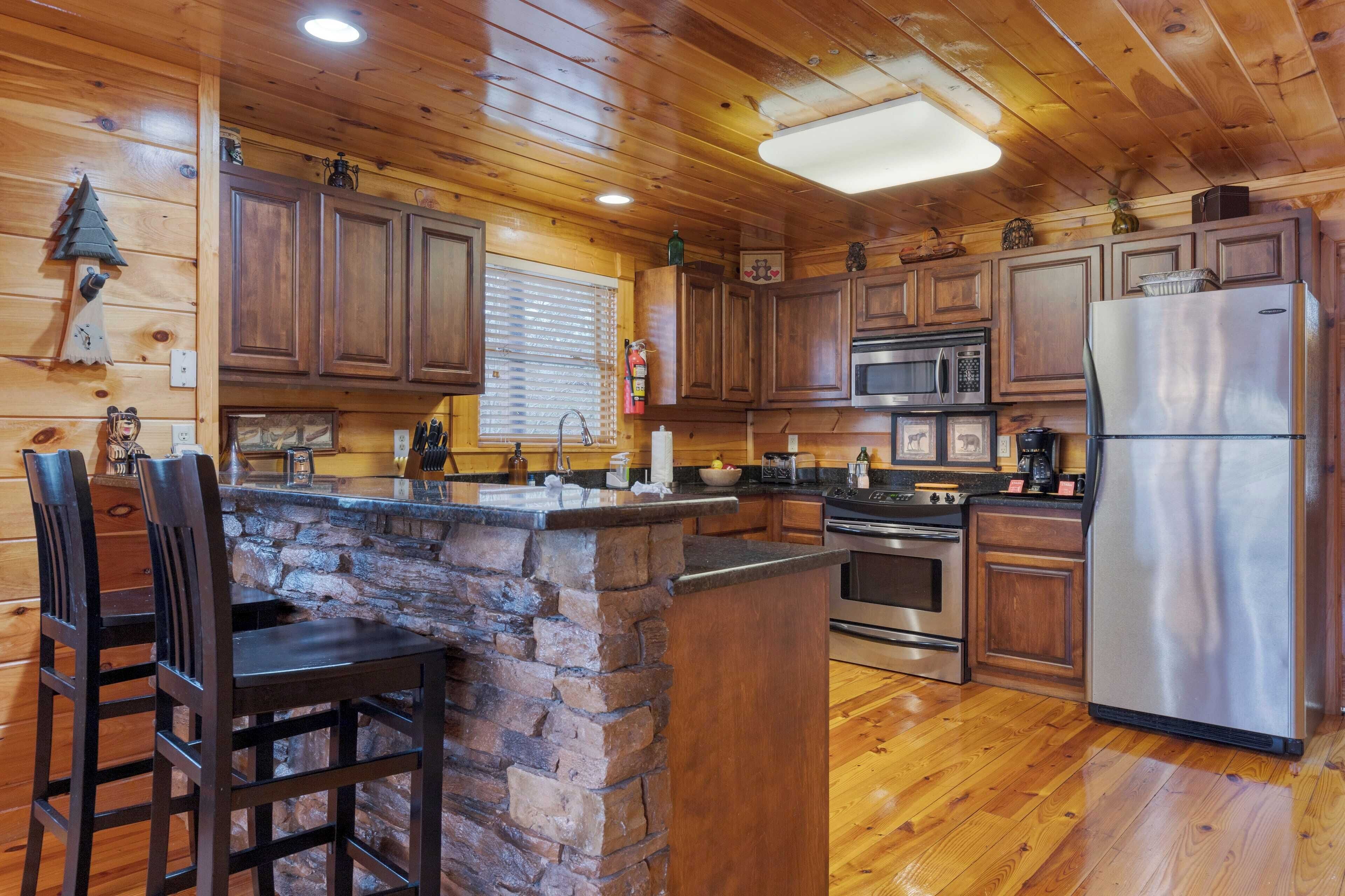 A wooden cabin interior with a stone island bar, wooden cabinets, a stainless steel refrigerator, and a stone fireplace.