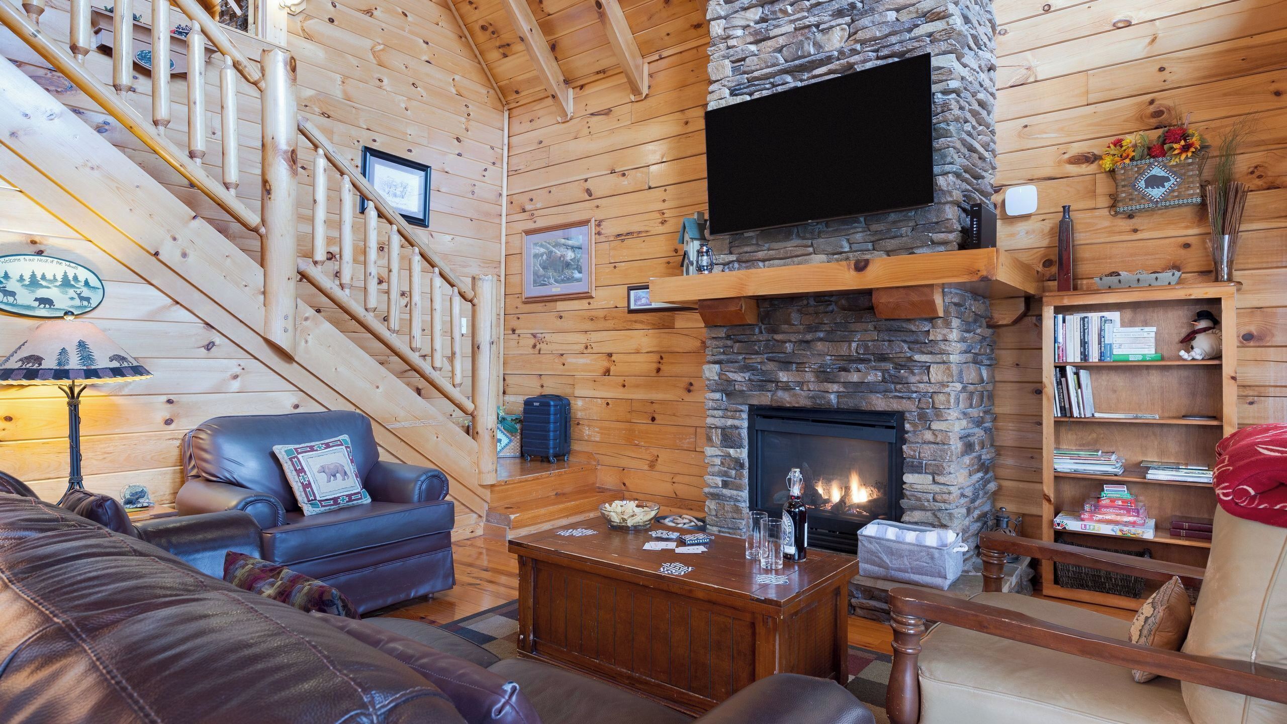 Ein Holzblockhaus-Interieur mit Kamin aus Stein, einem an der Wand befestigten Fernseher, einer Treppe und einem Ledersofa.