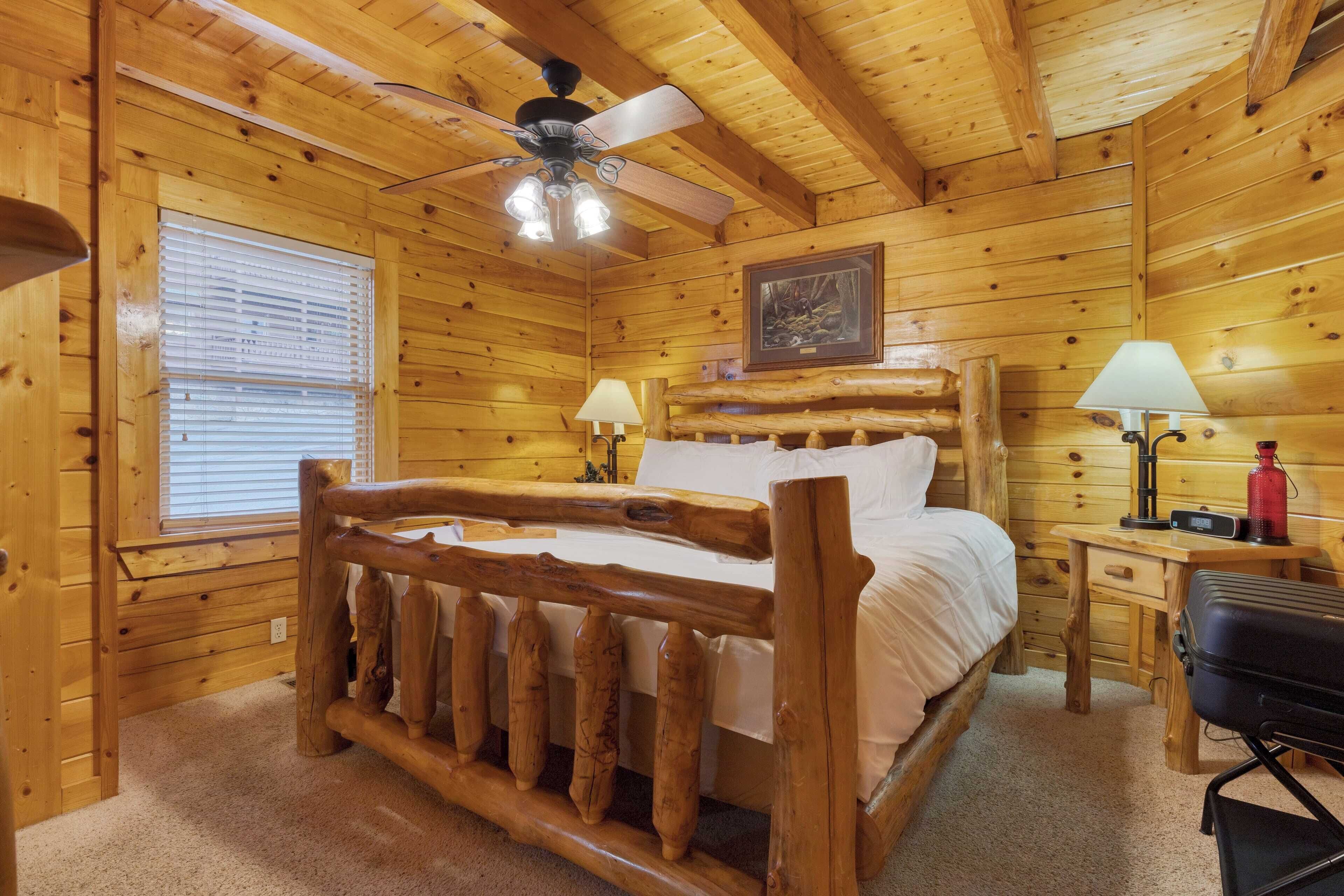 Una habitación de cabaña de madera con una cama, dos mesitas de noche, un escritorio, una silla, un ventilador de techo y una ventana con persianas.