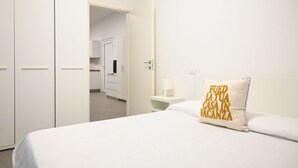 A hotel room with a bed, a nightstand with a lamp, a white door, and a hallway leading to a bathroom. - SE136 – Senigallia, brand new two-room apartment on the ground floor (Senigallia)