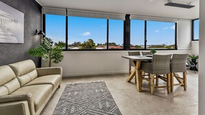 A modern living room with a beige sofa, dining table with chairs, large windows, and a potted plant. - Luxury accommodation close to the beach and main street. (Kingston SE)