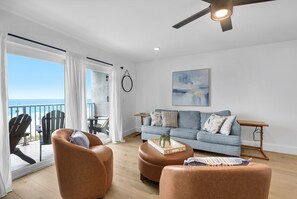 A modern living room with a sofa, ottoman, and chairs, a ceiling fan, and a view of the sea through large windows.
