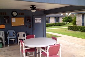 Un espace extérieur aménagé avec une table et des chaises, protégé par un auvent, équipé d’un ventilateur de plafond, d’une horloge et d’un panneau indiquant qu’il est réservé aux personnes de 55 ans et plus.