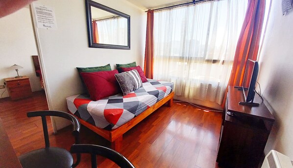 Una habitación acogedora con piso de madera, una cama con manta a cuadros, una silla, un escritorio con computadora y una ventana con cortinas.