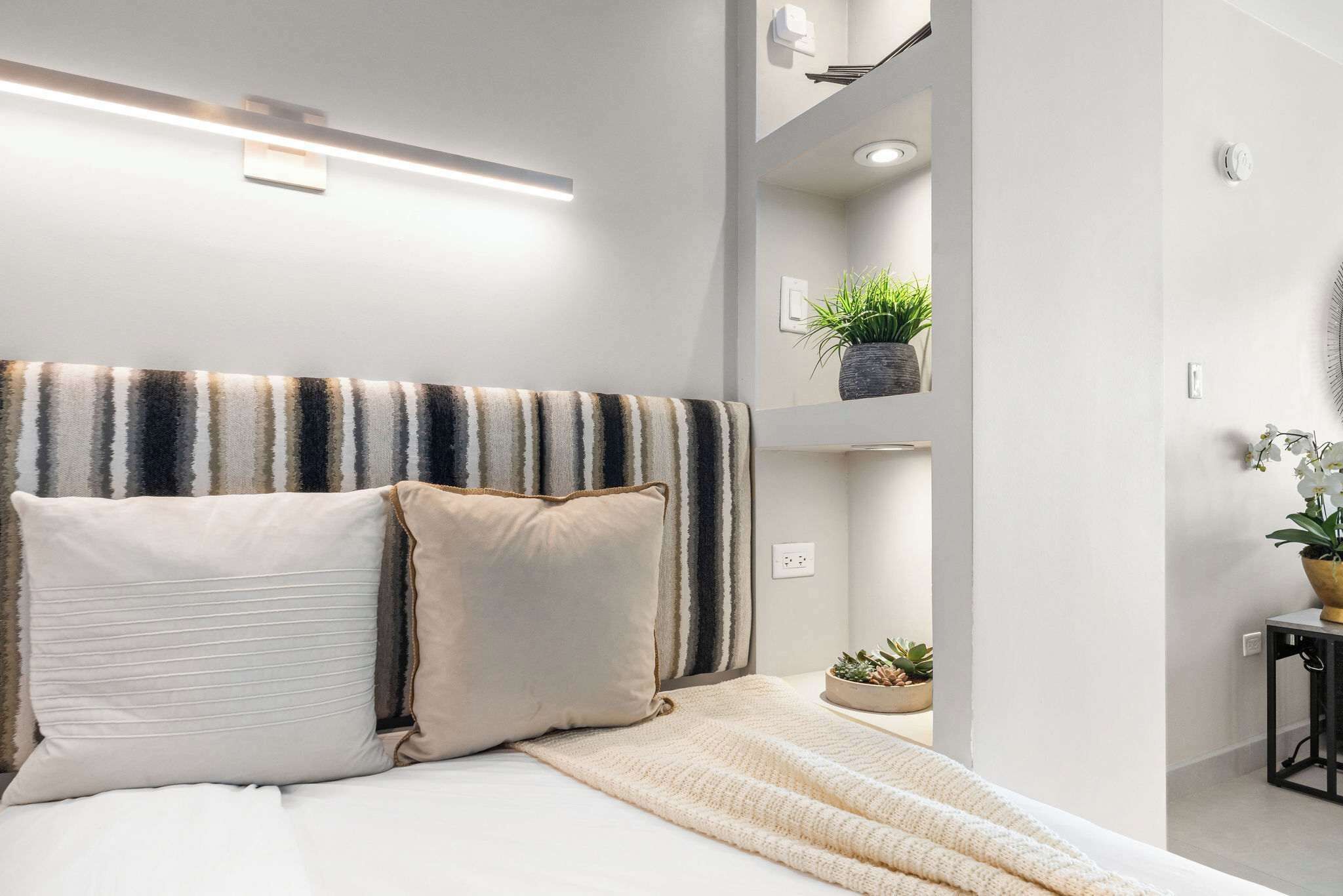 A modern bedroom with a striped headboard, a bedside table with a plant, and a wall-mounted shelf with a plant.