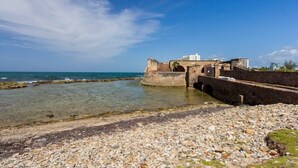 A coastal area with a rocky beach, a historic fort, and a modern building in the background. - Aqua Vista | Modern & Waterfront in Best Location (San Juan)