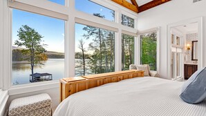A bedroom with large windows, a wooden bench, and a view of a lake and forest. - New Designer Lakefront Dock Putting Green Golf Sim (Nebo)