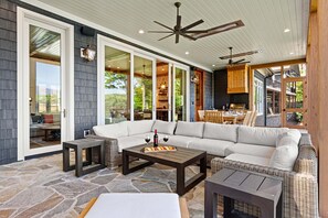 A covered patio area with a sectional sofa, a wooden table, and a stone floor. - New Designer Lakefront Dock Putting Green Golf Sim (Nebo)