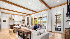 A spacious living area with a large sofa, wooden dining table, and a kitchen in the background. - New Designer Lakefront Dock Putting Green Golf Sim (Nebo)