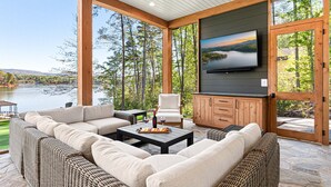 A spacious outdoor living area with a large sofa, a dining table, and a television mounted on the wall. - New Designer Lakefront Dock Putting Green Golf Sim (Nebo)