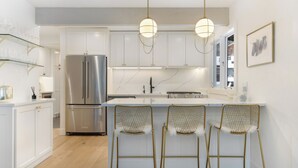 A modern kitchen with white cabinetry, a stainless steel refrigerator, a marble countertop, and three bar stools with gold frames.