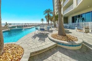 Un área de piscina con sillones de descanso y un jacuzzi, rodeada de palmeras y un edificio con balcones.