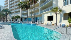 A swimming pool with lounge chairs and palm trees, surrounded by a multi-story building with balconies. - Admiral Quarters 705 | Beachfront | Indoor & Outdoor Pools | Beach Getaways (Orange Beach)