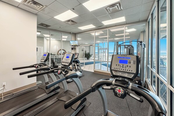Un gimnasio con cintas de correr, equipamiento de ejercicio y amplias ventanas con vistas.