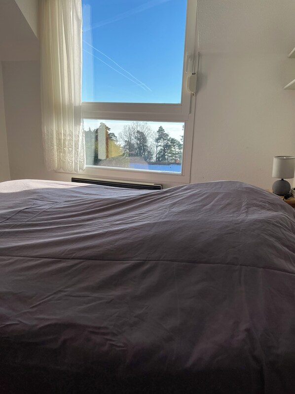 Une chambre avec une grande fenêtre, un lit recouvert d’une couette blanche et une lampe de chevet.