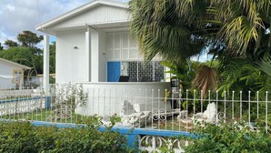 A white building with a blue door, surrounded by a white fence and lush greenery. - Spacious 6-bedroom house in lovely Christ Church with WiFi, AC (Christ Church)
