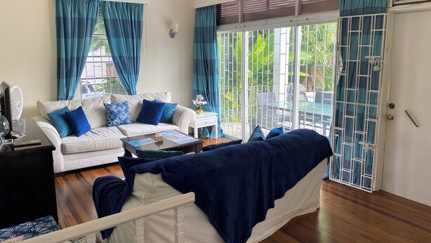 A modern living room with a white sofa, blue cushions, a coffee table, and a view through a glass door to an outdoor area with greenery. - Spacious 6-bedroom house in lovely Christ Church with WiFi, AC (Christ Church)
