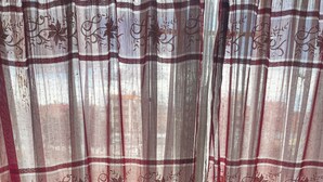 A room with a window covered by sheer curtains with floral patterns, a knitted throw on a chair, and a dark floor. - Beautiful house for rent in front of the Illimani in the city of La Paz.  (El Alto)