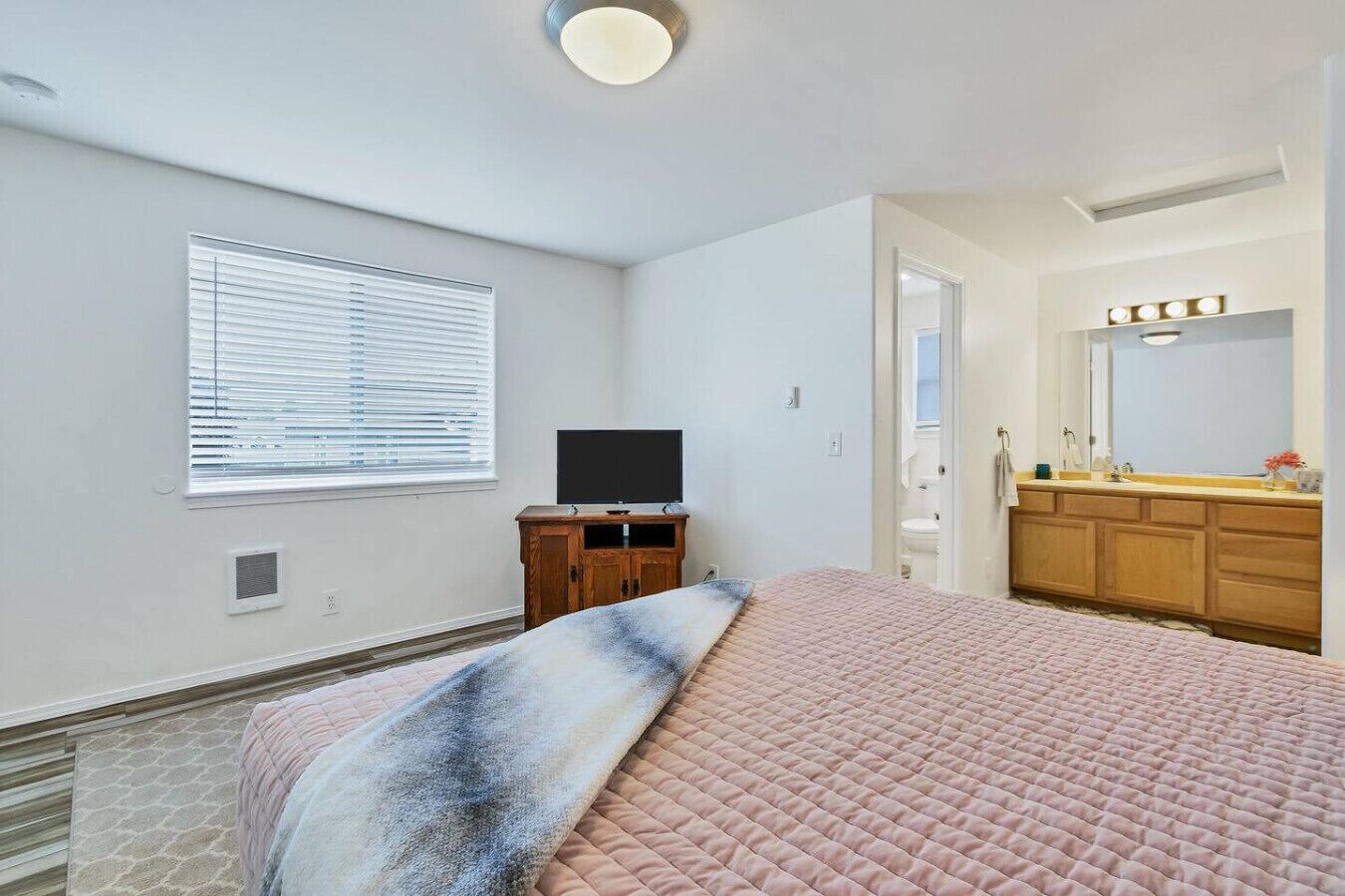 A bedroom with a bed, a television on a wooden stand, a bathroom with a vanity and mirror, and a window with blinds.