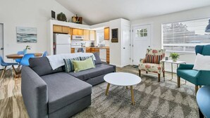 Een moderne woonkamer met een grijze bank, een ronde salontafel en een teal fauteuil. In de achtergrond is een kleine keuken te zien met houten kastjes en een magnetron.