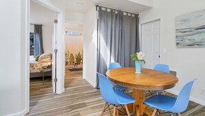 Een eetruimte met een ronde houten tafel en blauwe stoelen, een vaas met bloemen en uitzicht op een slaapkamer met een bed en gordijnen.