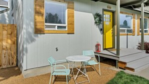 A single-story building with a porch, a yellow door, and a small outdoor seating area with two chairs and a table.