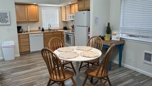 A compact kitchen with wooden cabinets, a dining table with four chairs, and a window with blinds.