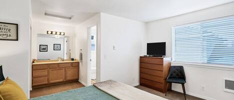 A bedroom with a bed, dresser, TV, and a window with blinds.