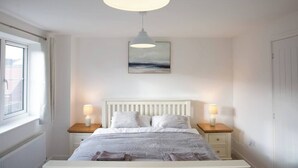 A bedroom with a bed, two bedside tables, a hanging light fixture, a window, and a wall-mounted painting.