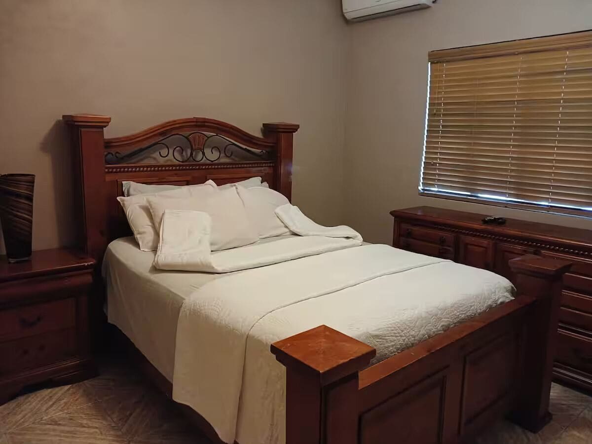 A neatly made bed with a wooden headboard, a bedside table, and a window with blinds.