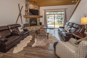 Una sala de estar acogedora con sofás de cuero, una chimenea de piedra y un ventanal con vistas a árboles cubiertos de nieve.