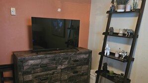 A room with a TV on a wooden dresser, a bed with a beige blanket, and a shelf with decorative items.