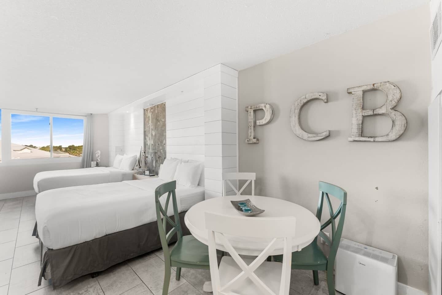 Habitación de hotel con dos camas, una mesa redonda y sillas. En la pared se ven letras grandes que dicen 'PCB'.