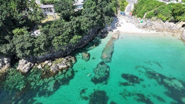 An aerial view of a coastal area with clear turquoise waters, rocky formations, and a beach.