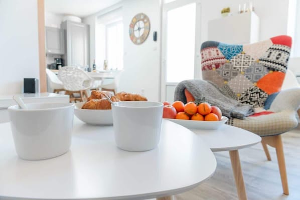 A modern living room with a round white table, two white mugs, a bowl of oranges, and a plate of croissants.
