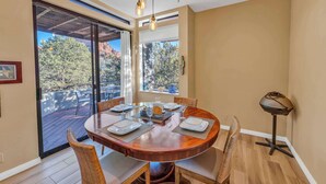 A dining area with a round table set for four, surrounded by wicker chairs, a wooden floor, and a view of trees and mountains through large glass doors.
