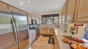 A modern kitchen with stainless steel appliances, wooden cabinets, and a large window offering a view of trees.