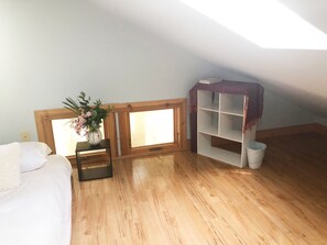 Ein Zimmer mit Holzboden, einem Bett mit weißer Bettwäsche, einem kleinen Tisch mit Blumenvase, einem Holzschrank mit Regalen und einem Fenster mit Blick.