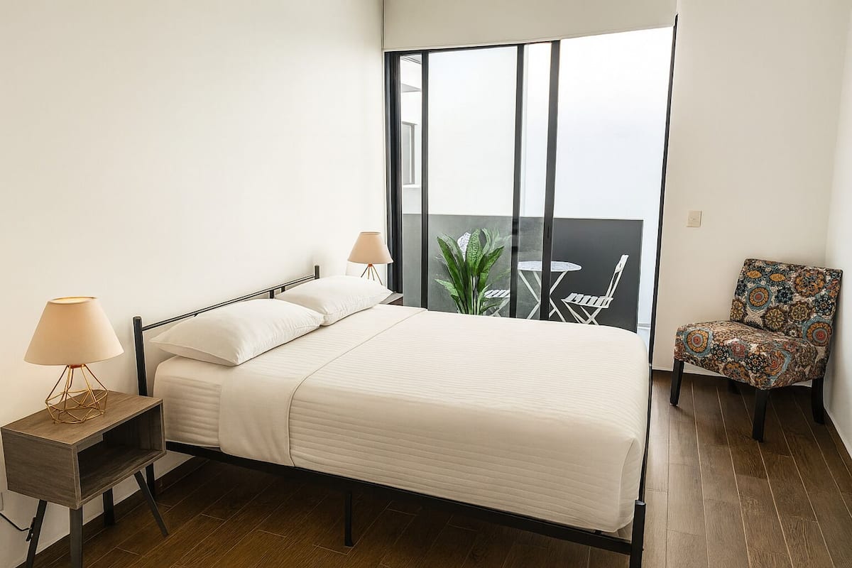 A bedroom with a bed, two bedside lamps, a chair, a balcony with a table and chairs, and a potted plant.