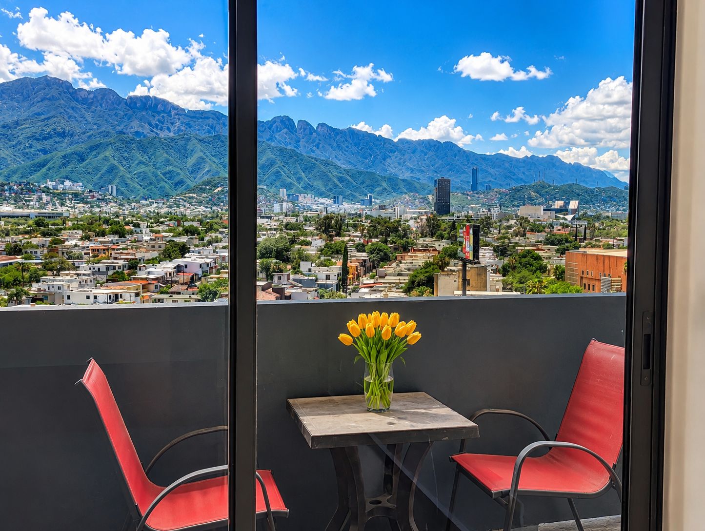 A balcony with a table and chairs, a vase with yellow flowers, and a cityscape with mountains in the background.