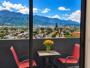 A balcony with a table and chairs, a vase with yellow flowers, and a cityscape with mountains in the background. - Panoramic Lofts La Silla (Monterrey)