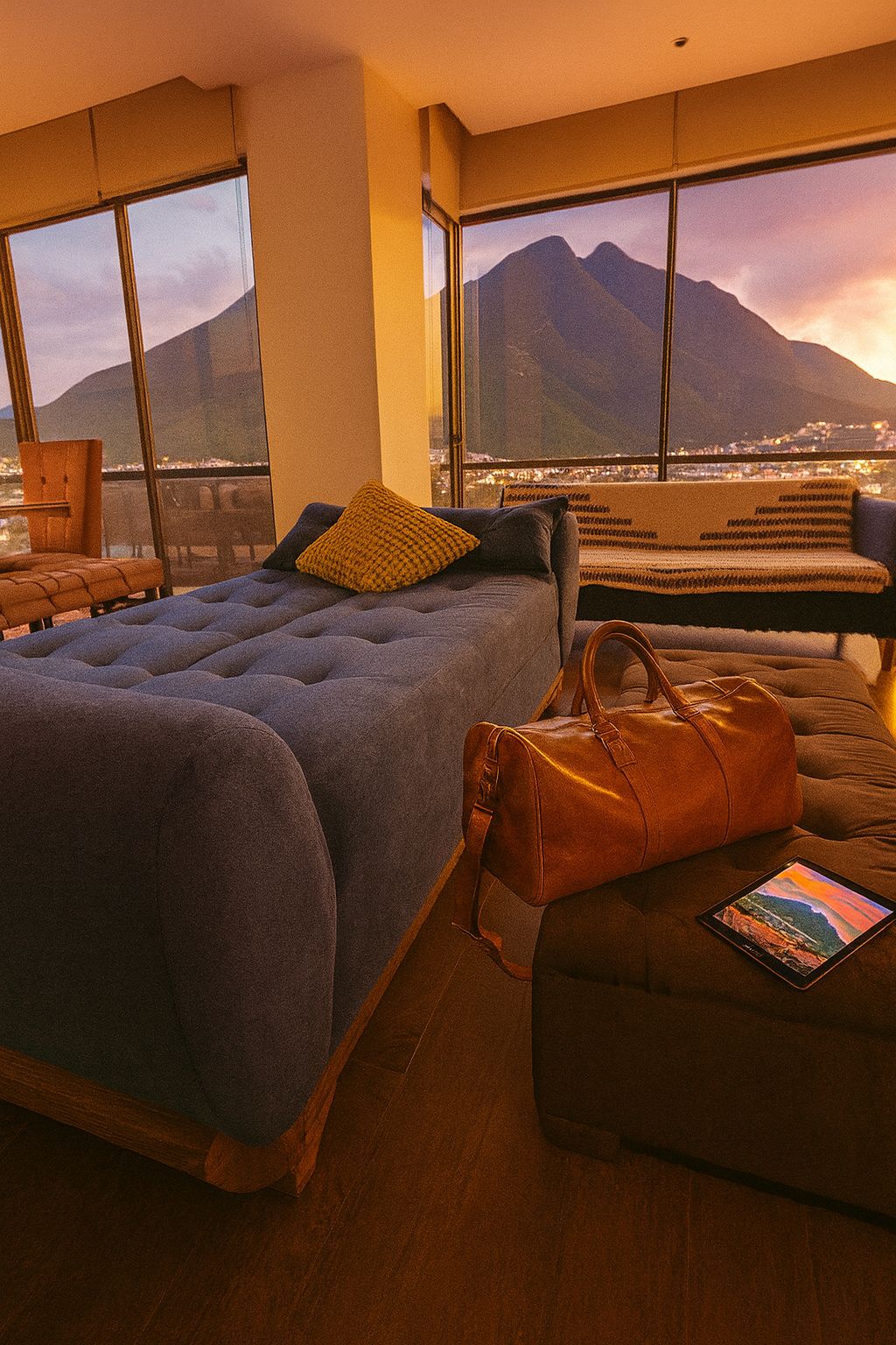 A bedroom with a bed, a sofa, a large window with a mountain view, and a leather bag on the sofa.