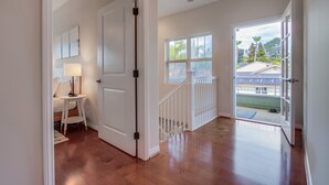 A hallway with wooden flooring, a white door, a small table with a lamp, and a view of a balcony with a railing. - Outdoor Living Area * A/C * Custom Built Home (Encinitas)