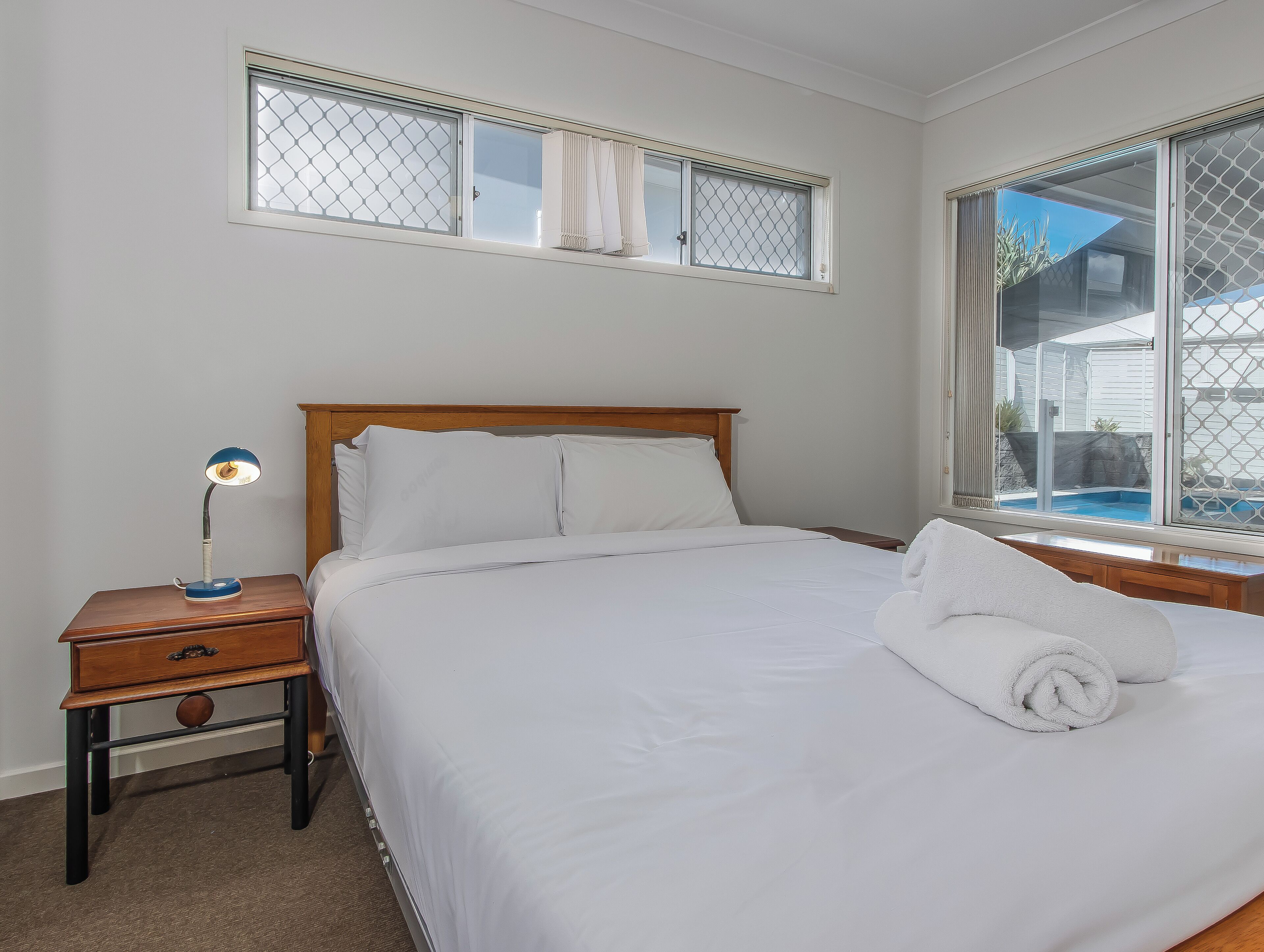 A neatly made bed with white linens, a wooden headboard, a bedside table with a lamp, and a window with a view of a pool and house.