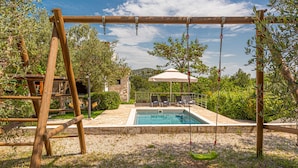 A wooden swing set, a pool, and a covered seating area with a view of the surrounding greenery.