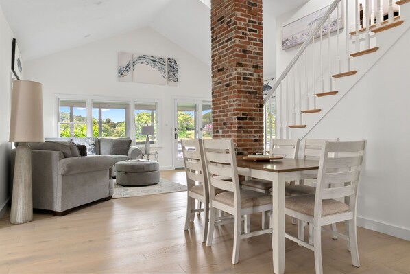 A spacious living room with a grey sofa, a dining table with white chairs, and a brick fireplace.